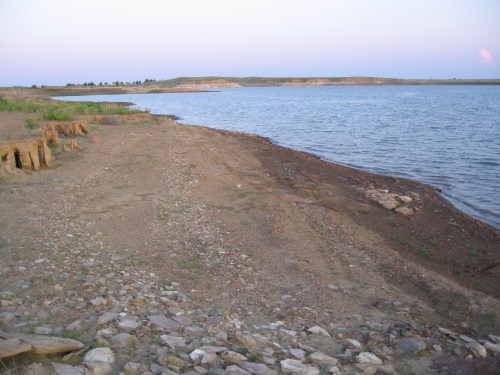 A serene lakeshore at dusk, featuring a rocky beach and gentle water, with grassy hills in the background.