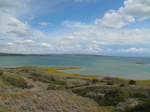 A scenic view of a calm lake surrounded by grassy hills and a cloudy blue sky.