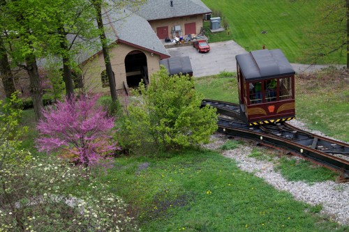 A small train on a track near a house, surrounded by green grass and blooming trees in a scenic outdoor setting.