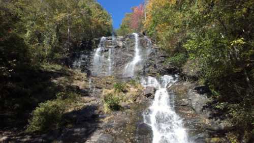 A scenic waterfall cascading down rocky cliffs, surrounded by lush greenery and autumn-colored trees under a clear blue sky.