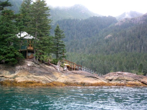 A rocky shoreline with a wooden cabin nestled among trees, surrounded by misty mountains and calm water.