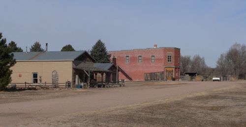 A dusty road leads to a small, rustic town with a mix of wooden and brick buildings under a clear blue sky.