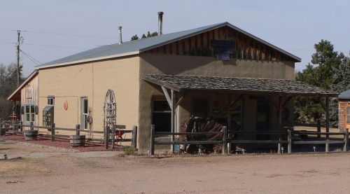 A rustic building with a wooden porch, surrounded by a dirt area and wooden fencing, set against a clear sky.
