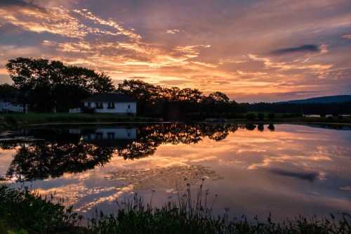 A serene sunset over a calm lake, reflecting colorful clouds and trees, with a white house in the foreground.