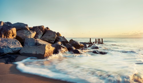 Waves gently wash over rocks at the shore, with wooden posts emerging from the water under a soft sunset glow.