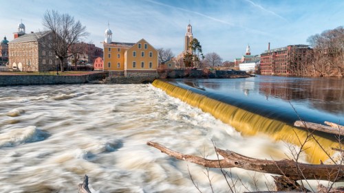 A river with a waterfall flows past historic buildings and trees under a clear blue sky.