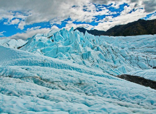 A vast glacier with jagged ice formations under a cloudy blue sky and distant mountains.