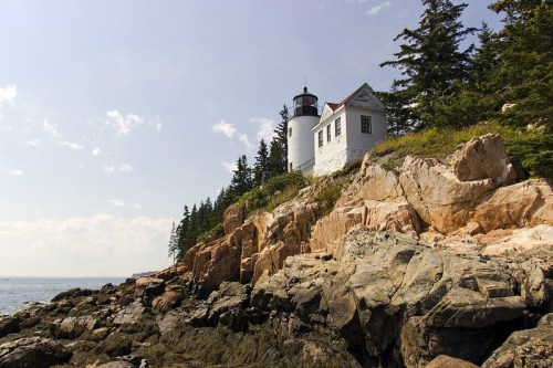A white lighthouse stands on rocky cliffs, surrounded by trees, overlooking the ocean under a clear blue sky.