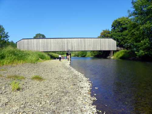 A wooden bridge spans a river, with people walking along the shore and greenery in the background under a clear blue sky.