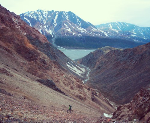 A hiker stands in a rocky valley surrounded by mountains and a distant lake under a cloudy sky.