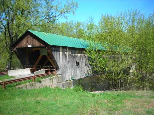 A rustic covered bridge with a green roof, surrounded by trees and grass, spanning a calm waterway.