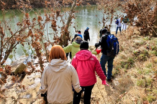 A group of people walking along a riverbank, surrounded by trees and autumn foliage.