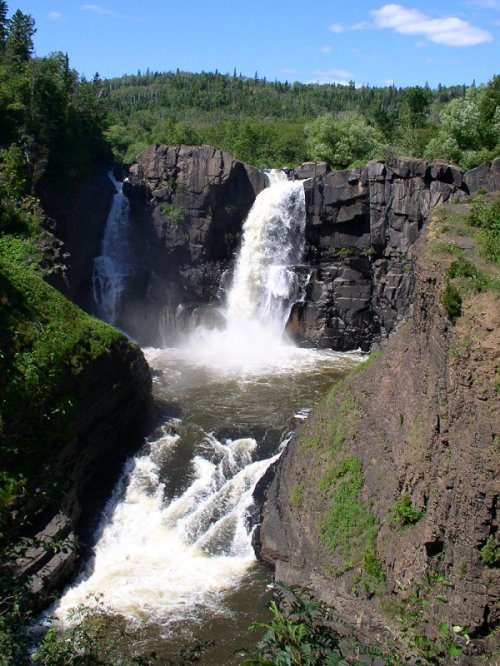 A scenic waterfall cascading down rocky cliffs, surrounded by lush greenery and a clear blue sky.