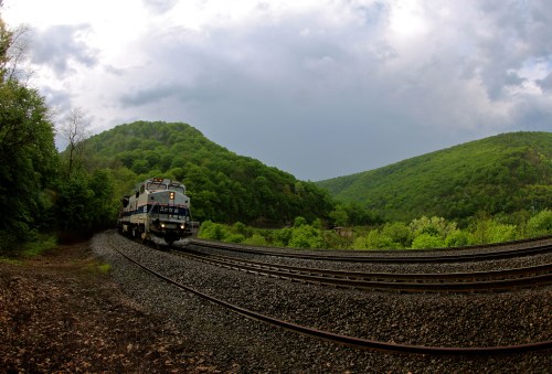 A train navigates curved tracks surrounded by lush green mountains under a cloudy sky.