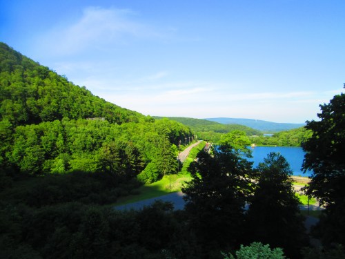 Lush green hills surround a serene lake under a clear blue sky, with a winding road visible in the foreground.