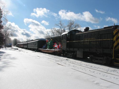 A vintage train in a snowy landscape, decorated with festive items and surrounded by trees under a bright blue sky.