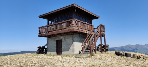 A wooden lookout tower on a rocky hill, with stairs leading up and a motorcycle parked nearby, against a clear blue sky.