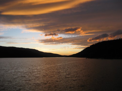 A serene lake at sunset, with vibrant clouds reflecting on the water and mountains in the background.