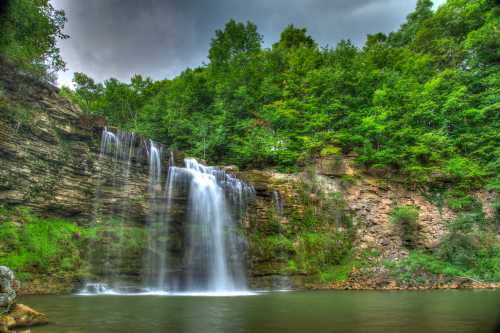 A serene waterfall cascades over rocky cliffs, surrounded by lush green trees and a calm pool below.