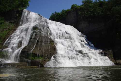 A cascading waterfall flows over rocky cliffs into a serene pool, surrounded by lush greenery under a clear blue sky.