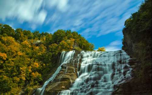 A majestic waterfall cascades down rocky cliffs, surrounded by vibrant autumn foliage and a blue sky.