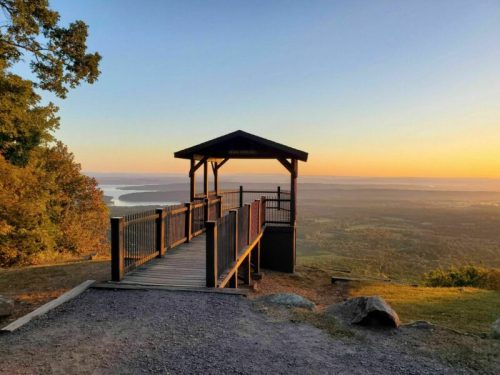 A wooden observation deck overlooks a valley at sunset, with a lake visible in the distance.