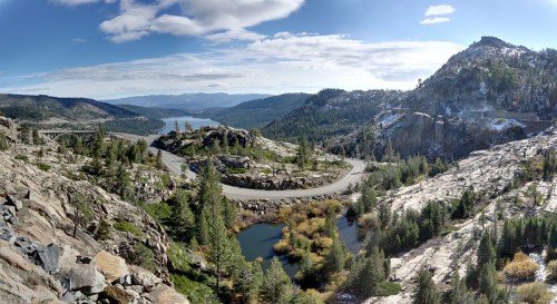 A panoramic view of rocky hills, winding roads, and a serene lake surrounded by trees under a clear blue sky.