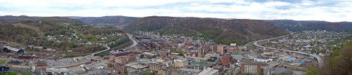 A panoramic view of a hilly town with buildings, roads, and green landscapes under a cloudy sky.