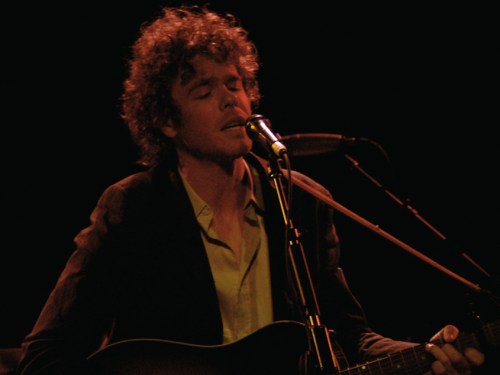 A musician with curly hair sings into a microphone while playing an acoustic guitar on stage.