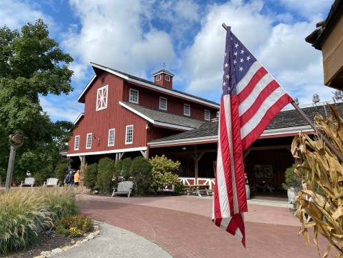 A red barn with a flag in front, surrounded by greenery and a pathway, under a partly cloudy sky.