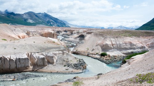 A winding river flows through a rugged, sandy landscape with mountains and cloudy skies in the background.