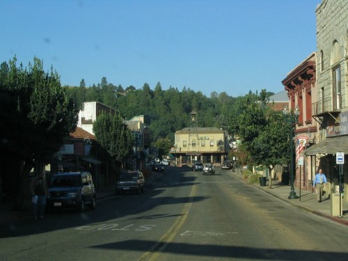A quiet street scene with shops and trees, set against a backdrop of hills under a clear blue sky.