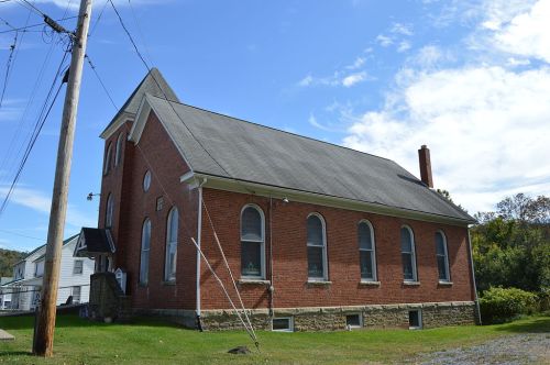 A red brick church with arched windows, set against a blue sky and surrounded by greenery and utility poles.