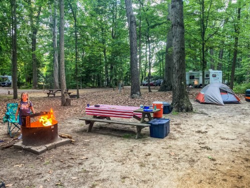 A campsite with a child by a fire pit, picnic table, cooler, and a tent surrounded by trees. A camper is in the background.