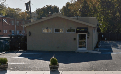 A small, modern restaurant building with a sign reading "KITCHEN" and a parking area in front. Trees in the background.