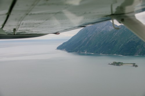 Aerial view of a coastline with mountains and a small island, seen from an airplane wing.
