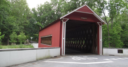 A red covered bridge with a wooden structure, surrounded by green trees, leading to a road with a stop sign.