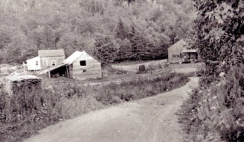 Black and white photo of a rural landscape with several old wooden buildings and a winding dirt road.