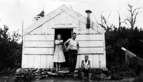 A black and white photo of three people outside a rustic cabin, with a sign reading "Glacierberry Camp" above the door.