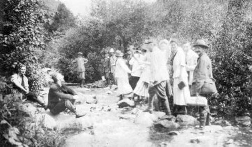 A group of people in early 20th century attire gathered by a stream, surrounded by trees and rocks.
