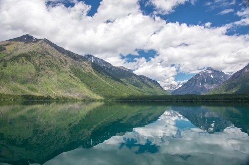 A serene lake reflecting lush green mountains and a cloudy blue sky.
