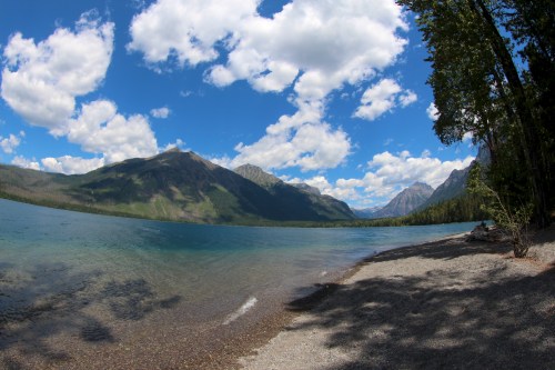 A serene lake surrounded by mountains under a blue sky with fluffy clouds, featuring a pebbly shoreline.