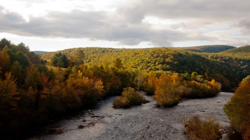 A serene river flows through a valley surrounded by colorful autumn foliage and rolling hills under a cloudy sky.