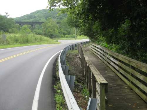 A winding road curves alongside a wooden walkway, surrounded by greenery and hills in the background.