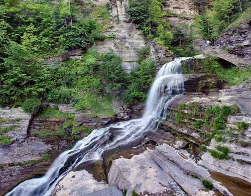 A serene waterfall cascades over rocky cliffs, surrounded by lush greenery and visitors enjoying the view.