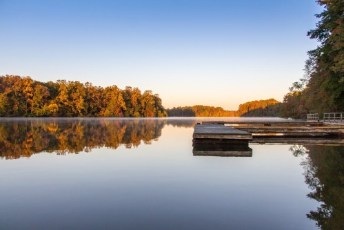 Serene lake at sunrise, with wooden docks and colorful autumn trees reflecting on the calm water.