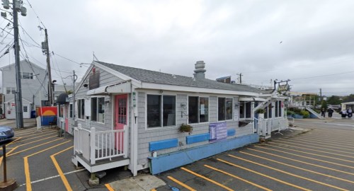 A small, charming white building with a gray roof, surrounded by parking spaces and power lines on a cloudy day.