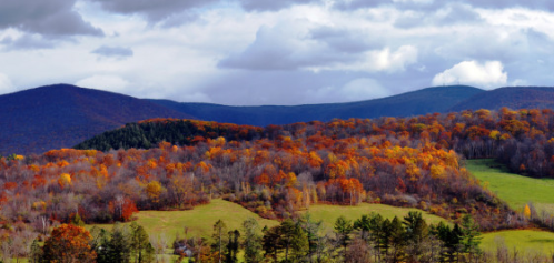 Vibrant autumn landscape with colorful trees, rolling hills, and a cloudy sky in the background.