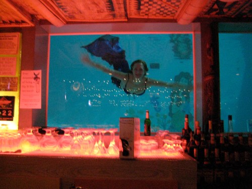 A mermaid swims gracefully behind a bar, illuminated by colorful lights in a vibrant underwater setting.