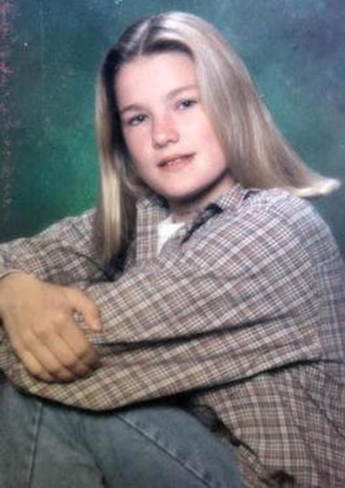 A young person with long hair sits with arms crossed, wearing a plaid shirt and jeans, against a colorful background.